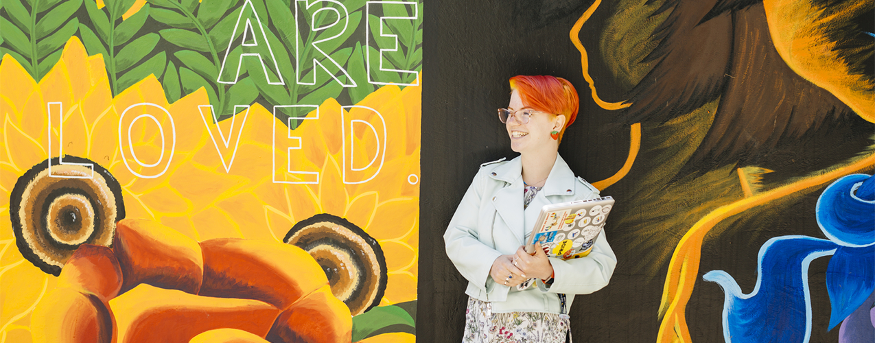 A UC Santa Cruz student stands in front of a colorful mural.